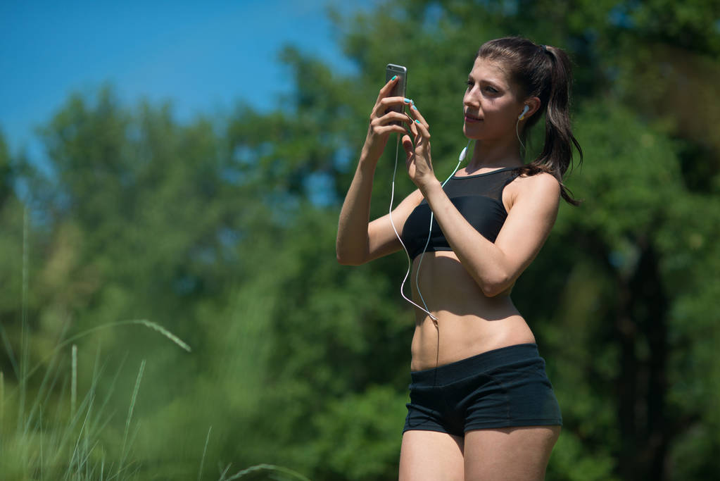 Jovem atleta procurando um smartphone ao ar livre. Ela escolhe a música para o teu treino.
 - Foto, Imagem