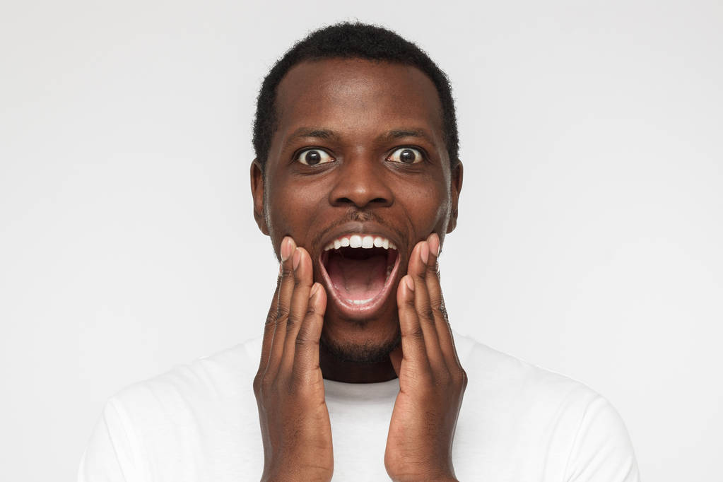 Lähikuva kuva nuori komea afrikkalainen amerikkalainen mies tyhjä valkoinen t-paita, eristetty valkoisella pohjalla, näyttää erittäin yllättynyt pyöreät silmät ja suu, positiivisesti järkyttynyt
 - Valokuva, kuva