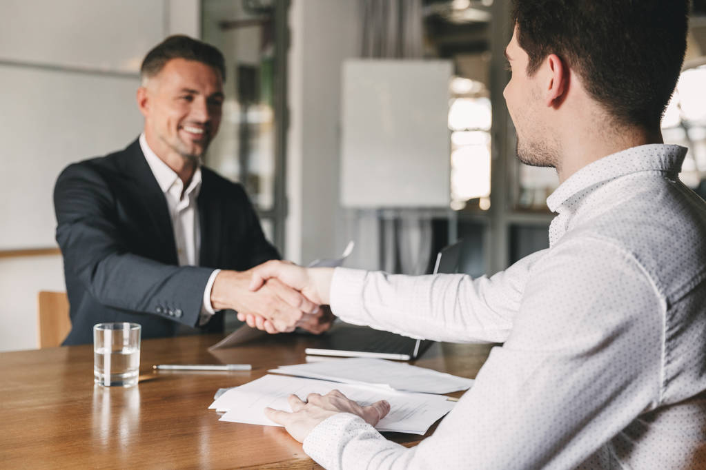 Бизнес, карьера и концепция - успешный молодой человек улыбается и пожимает руку европейскому бизнесмену после успешных переговоров или интервью в офисе
 - Фото, изображение