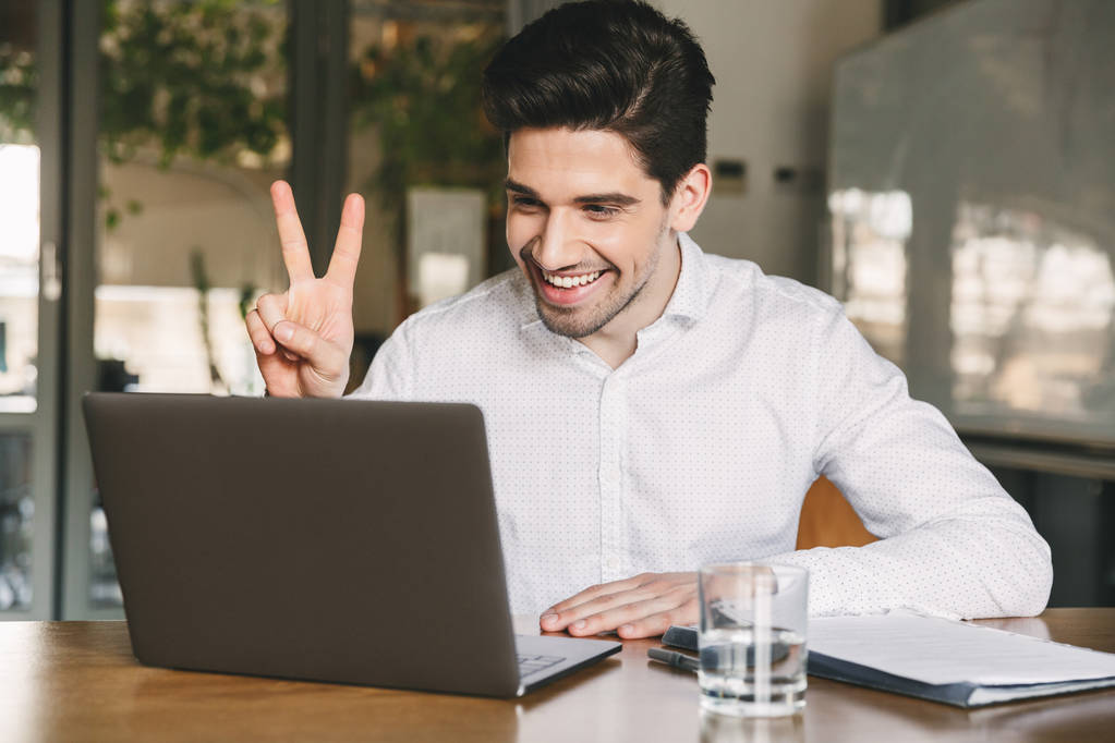 Imagen del alegre hombre de oficina de 30 años con camisa blanca riéndose y mostrando un signo de paz en la computadora portátil durante la videoconferencia o llamada
 - Foto, Imagen