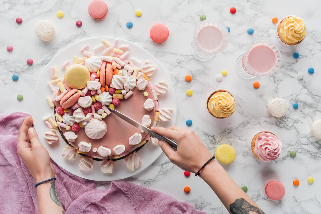 Ausgeschnittene Ansicht einer tätowierten Frau beim Anschneiden von rosa Marshmallow-Kuchen mit Messer auf dem Tisch  - Foto, Bild