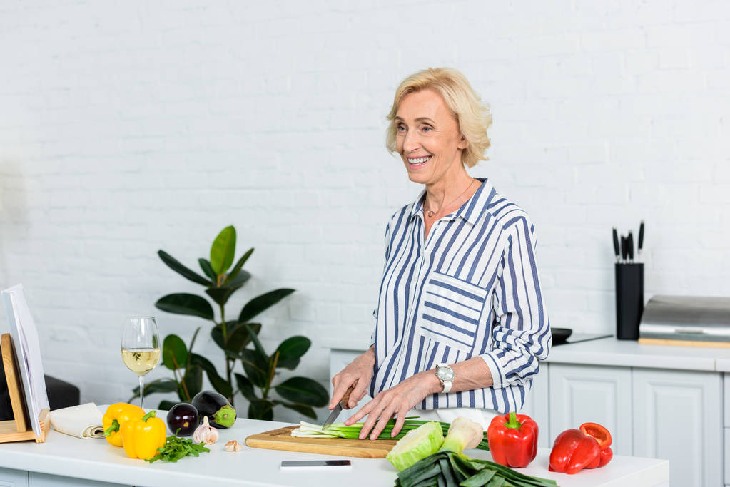 sonriente atractivo pelo gris mujer corte cebolla verde en la cocina
  - Foto, imagen