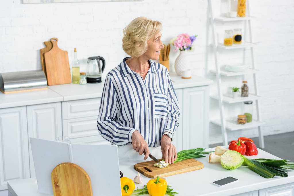 atractiva mujer de pelo gris cortando cebolla verde en la cocina y mirando hacia otro lado
 - Foto, imagen