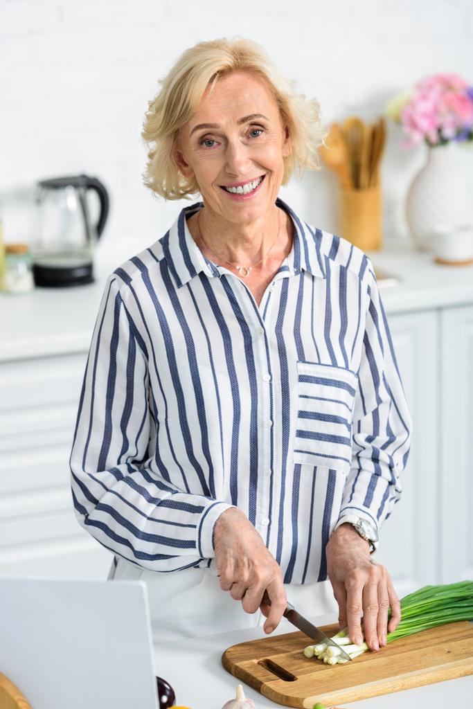 uśmiechnięta kobieta piękne siwe włosy cięcia zielona cebula w kuchni i patrząc na kamery - Zdjęcie, obraz