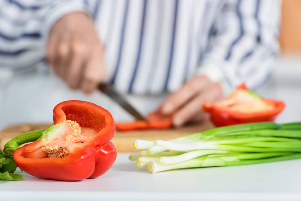 image recadrée d'une femme âgée coupant du poivron rouge sur une planche en bois dans la cuisine avec des légumes au premier plan
 - Photo, image