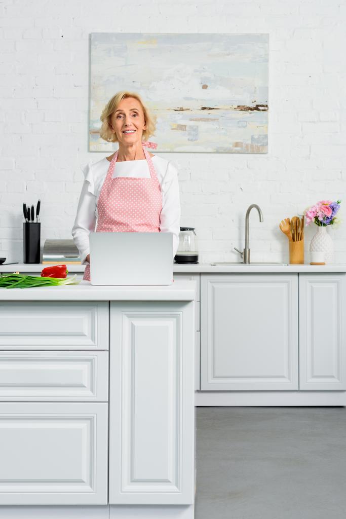 atrakcyjna kobieta starszego komputera przenośnego do gotowania w kuchni i patrząc na kamery - Zdjęcie, obraz