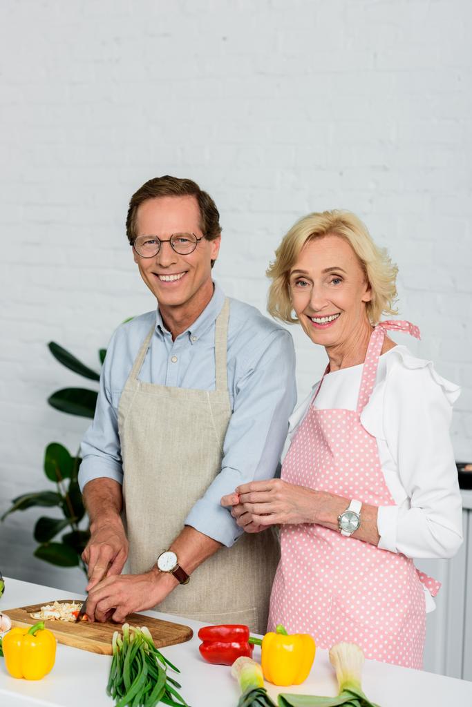 улыбающаяся пожилая пара готовит вместе на кухне и смотрит в камеру
 - Фото, изображение