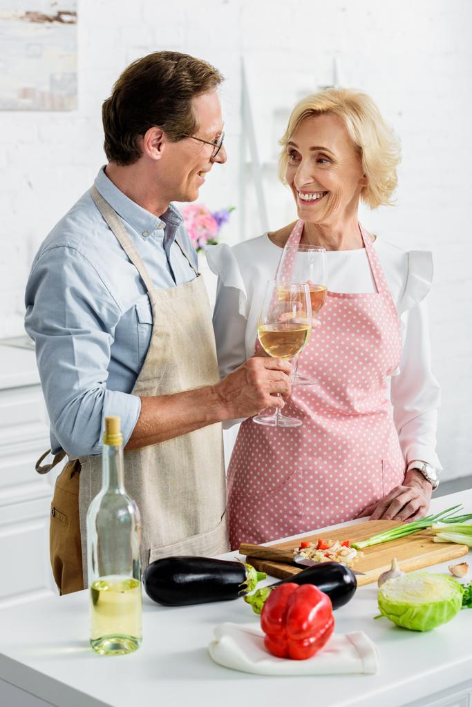 smiling senior couple clinking with glasses of wine during cooking at kitchen and looking at each other - Photo, Image