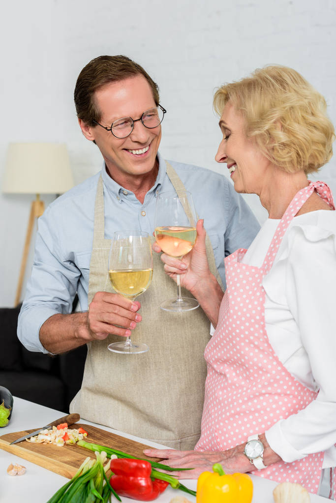 smiling senior couple clinking with glasses of wine during cooking at kitchen - Photo, Image