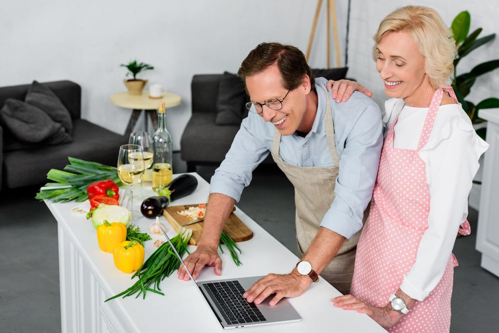 усміхнена старша пара готує на кухні і використовує ноутбук
 - Фото, зображення