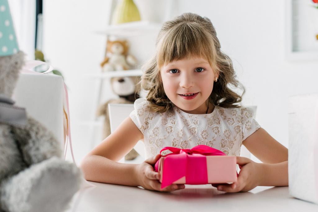 χαμογελαστό παιδί γενέθλια κρατώντας κουτί δώρου και βλέπουν φωτογραφική μηχανή στο τραπέζι  - Φωτογραφία, εικόνα