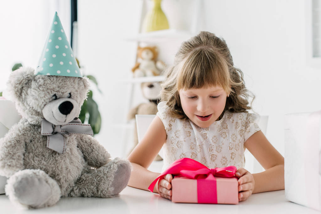 smiling birthday kid looking at gift box while sitting at table with teddy bear in cone - Photo, Image