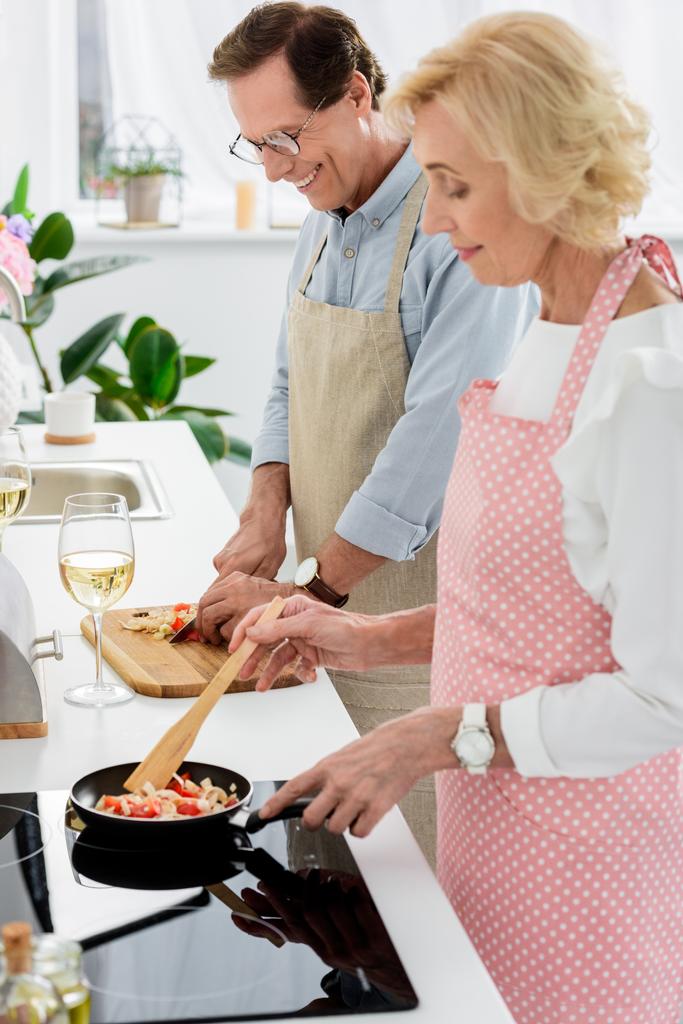 Zijaanzicht van senior paar koken van groenten op koekenpan en snijden groenten op houten bord op keuken - Foto, afbeelding