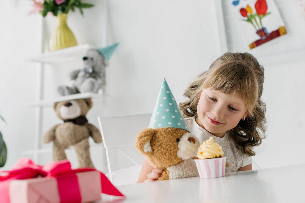 cute birthday kid feeding teddy bear in cone by cupcake at table  - Photo, Image