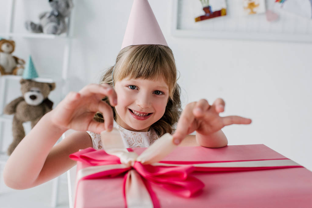 szczęśliwe dziecko w kształcie stożka, otwierając pudełko urodziny i patrząc na kamery - Zdjęcie, obraz