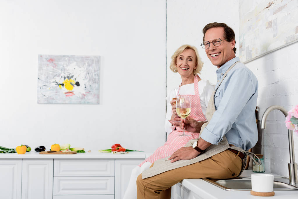 onnellinen vanhempi pari esiliinoissa pitäen viinilaseja ja hymyillen kameralle keittiössä
 - Valokuva, kuva