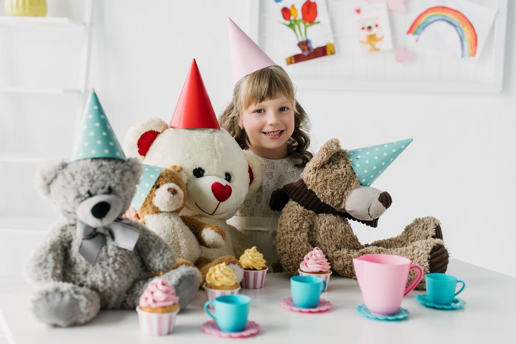 smiling birthday kid with teddy bears in cones having tea party with cupcakes at table  - Photo, Image