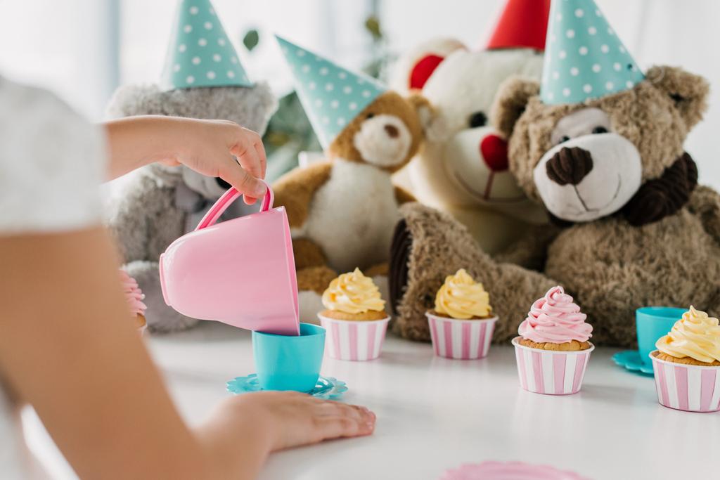 bijgesneden afbeelding van kid gieten thee in beker naar teddyberen in kegels aan tafel met cupcakes  - Foto, afbeelding