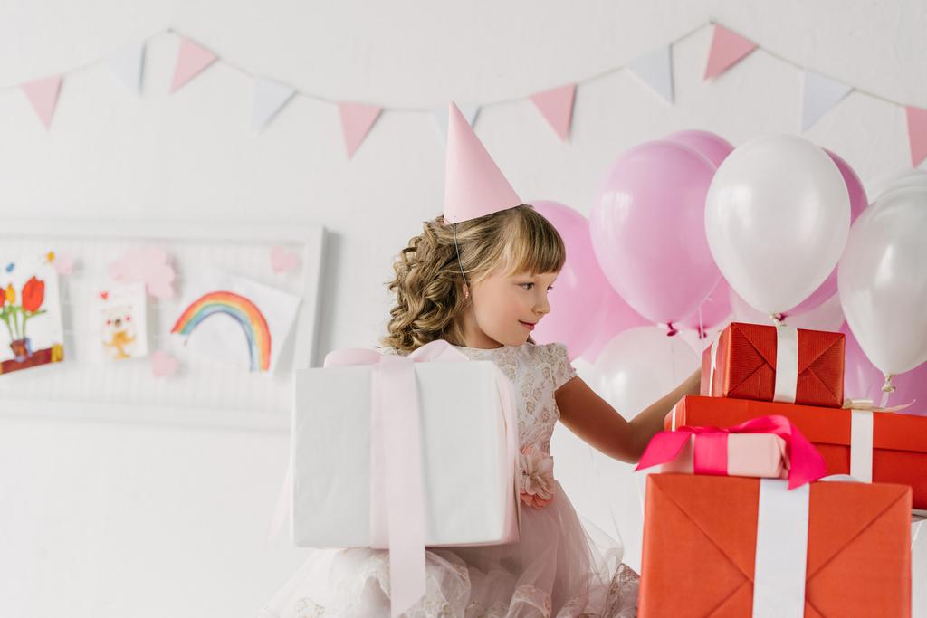 niño lindo cumpleaños en cono mirando cajas de regalo
  - Foto, Imagen