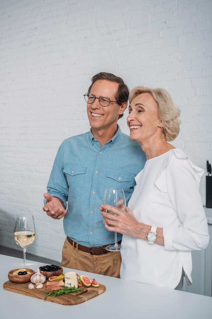 glückliches Seniorenpaar trinkt Wein und schaut weg, während es zu Hause leckere Snacks isst  - Foto, Bild