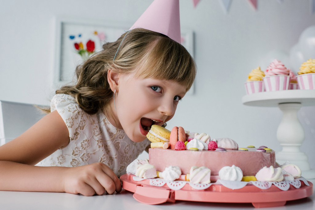χαμογελαστό μικρό παιδί σε κώνο τρώει τούρτα γενεθλίων και βλέπουν τα φωτογραφικών μηχανών - Φωτογραφία, εικόνα