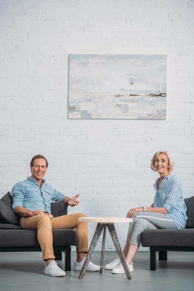 onnellinen vanhempi pari hymyilee kameralle istuessaan yhdessä kotona
  - Valokuva, kuva