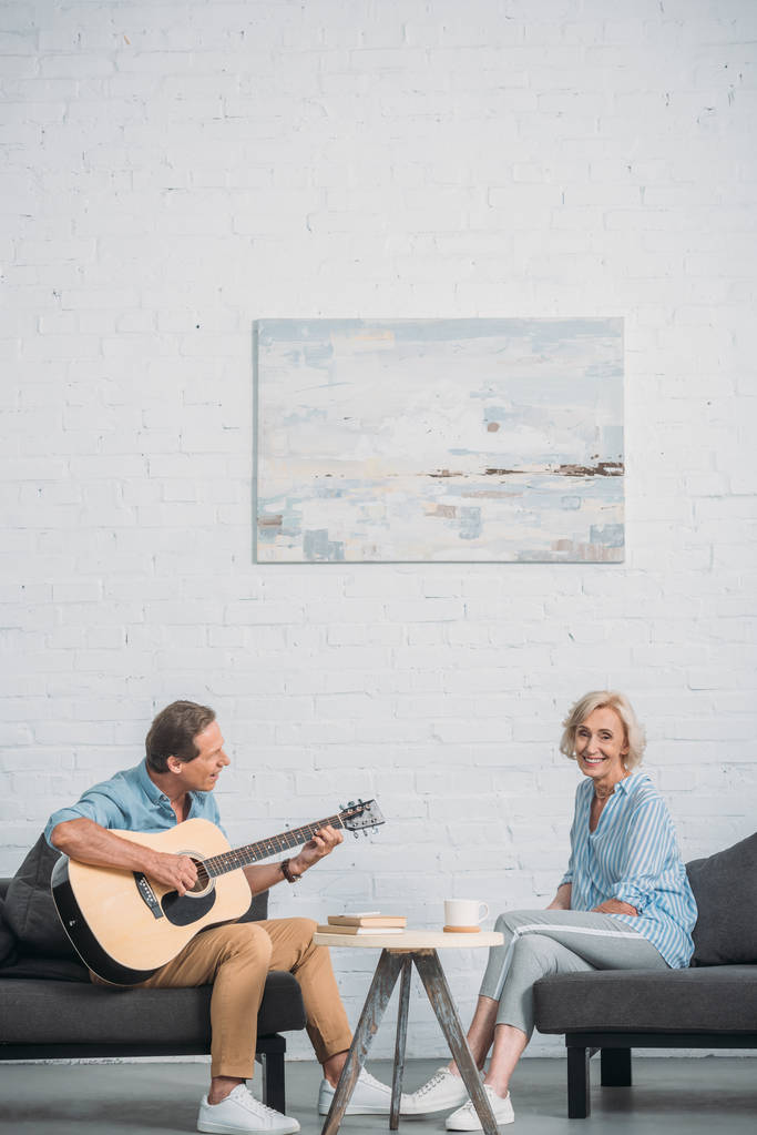 happy man playing acoustic guitar and looking at senior wife smiling at camera at home - Photo, Image