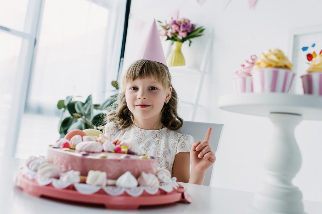 happy child in cone doing idea gesture at table with birthday cake  - Photo, Image