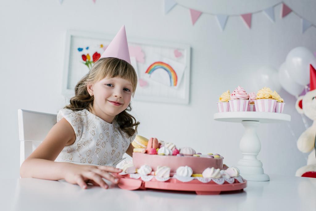 ευτυχισμένη αξιολάτρευτο παιδί σε κώνο που κάθεται στο τραπέζι με τούρτα γενεθλίων  - Φωτογραφία, εικόνα