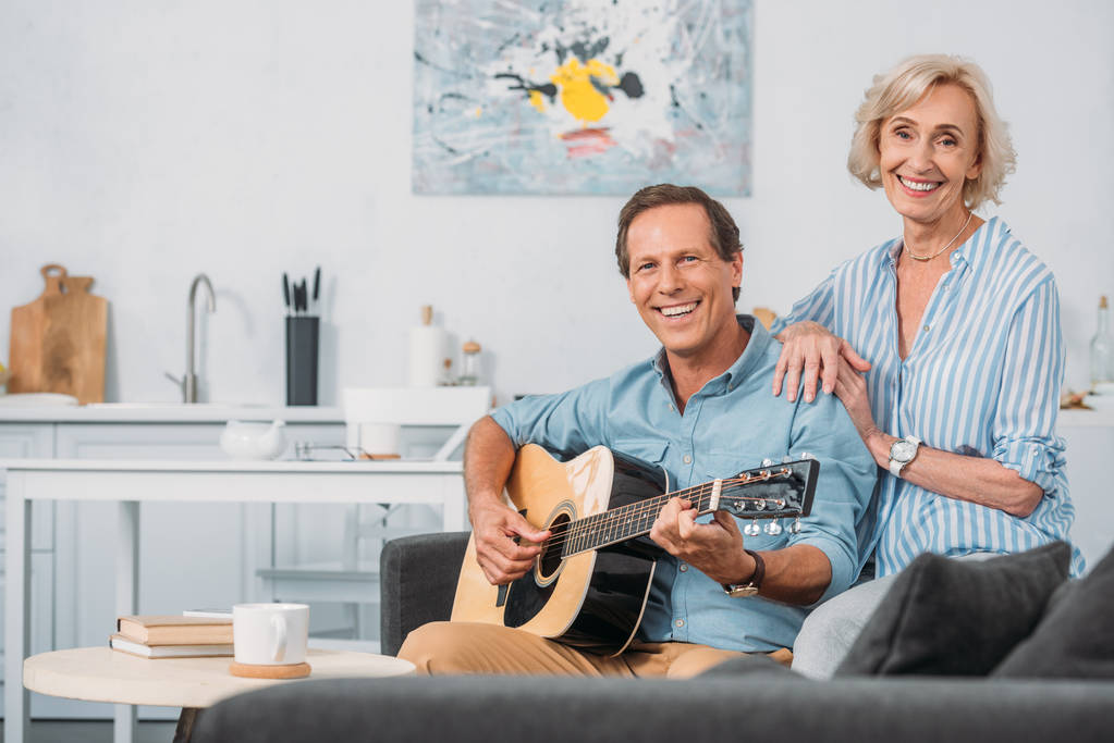 happy senior couple smiling at camera while man playing guitar at home - Photo, Image
