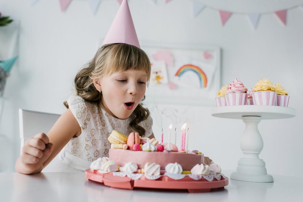 adorable niño en cono soplando velas de pastel de cumpleaños en la mesa
  - Foto, Imagen