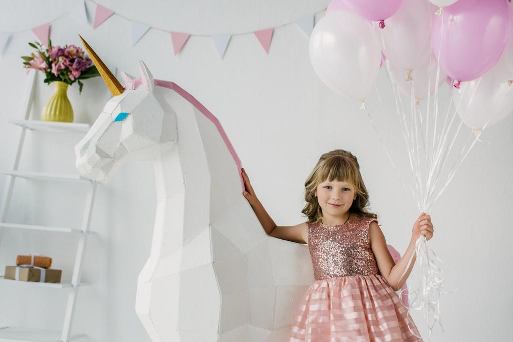 adorable little kid holding bunch of air balloons and standing with decorative unicorn  - Photo, Image