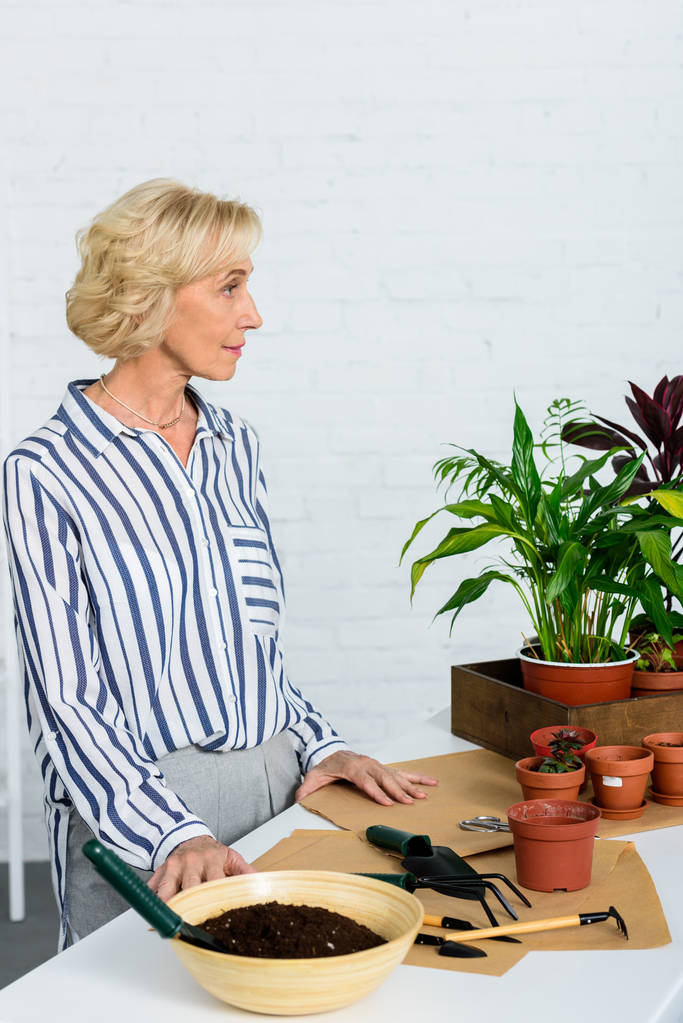 pensive senior woman looking away while cultivating potted plants at home - Photo, Image