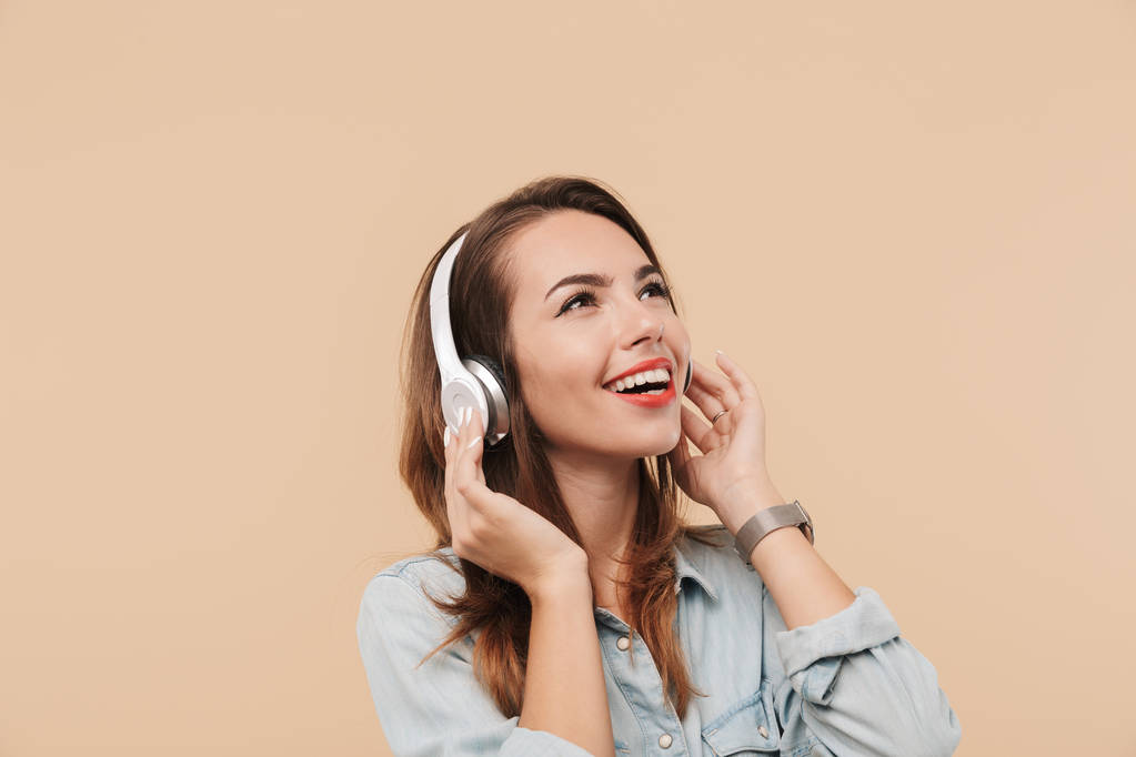 Imagen de una joven mujer feliz en auriculares escuchando música aislada sobre una pared de fondo beige
. - Foto, Imagen