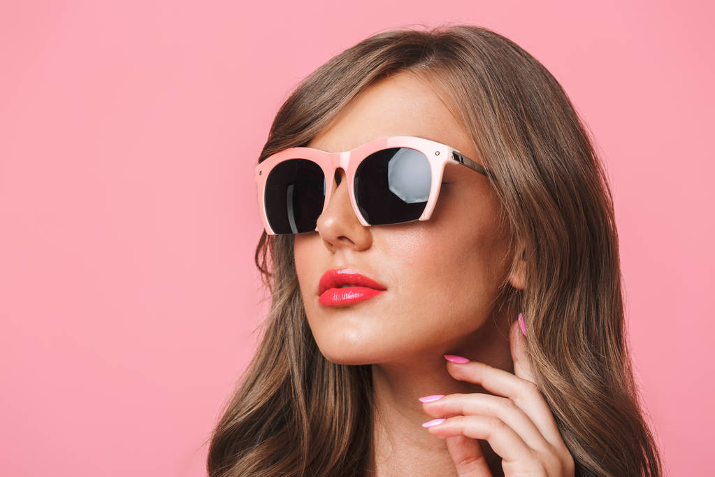 Beeld close-up van jonge Europese vrouw 20s met lang krullend kapsel dragen van een trendy zonnebril kijkt opzij met mode kijken geïsoleerd op roze achtergrond - Foto, afbeelding