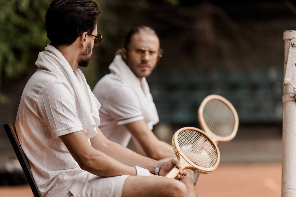 серьезные теннисисты в стиле ретро, сидящие на стульях с полотенцами и ракетками на теннисном корте
  - Фото, изображение