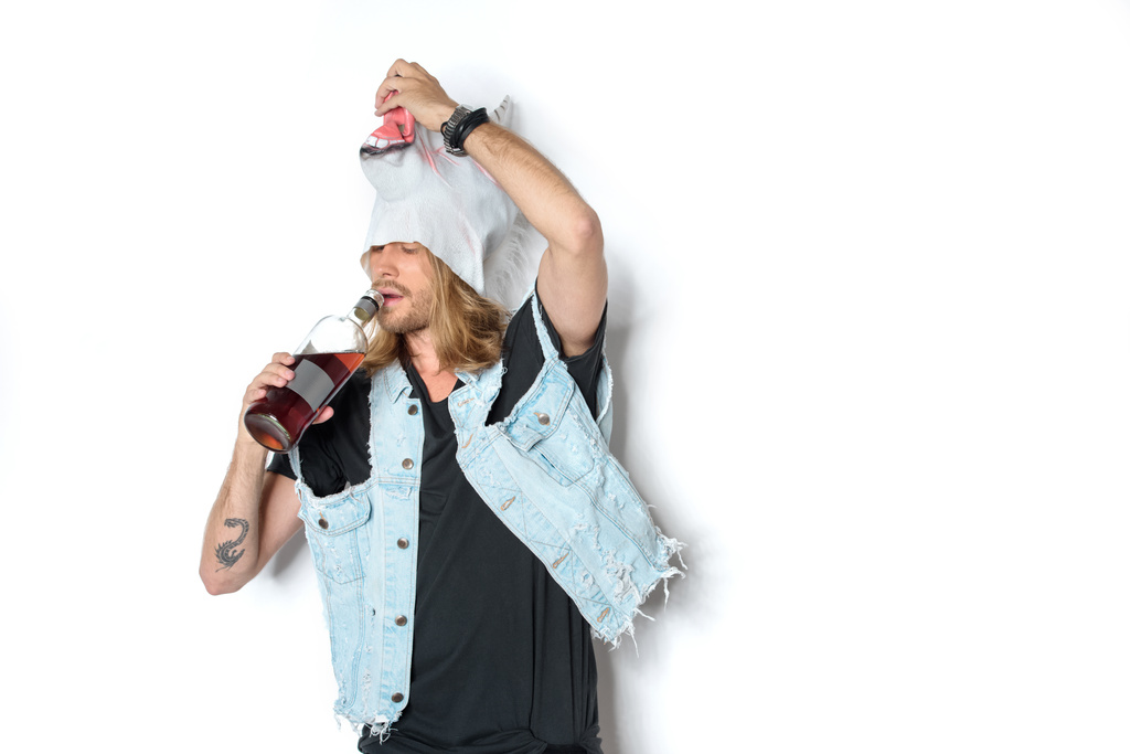 junger Mann in Einhornmaske und Jeansweste mit Hebemaske und trinkt Rum auf Weiß - Foto, Bild