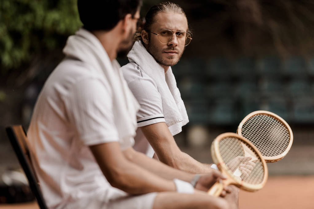 теннисисты в стиле ретро, отдыхающие на стульях с полотенцами и ракетками на теннисном корте
  - Фото, изображение