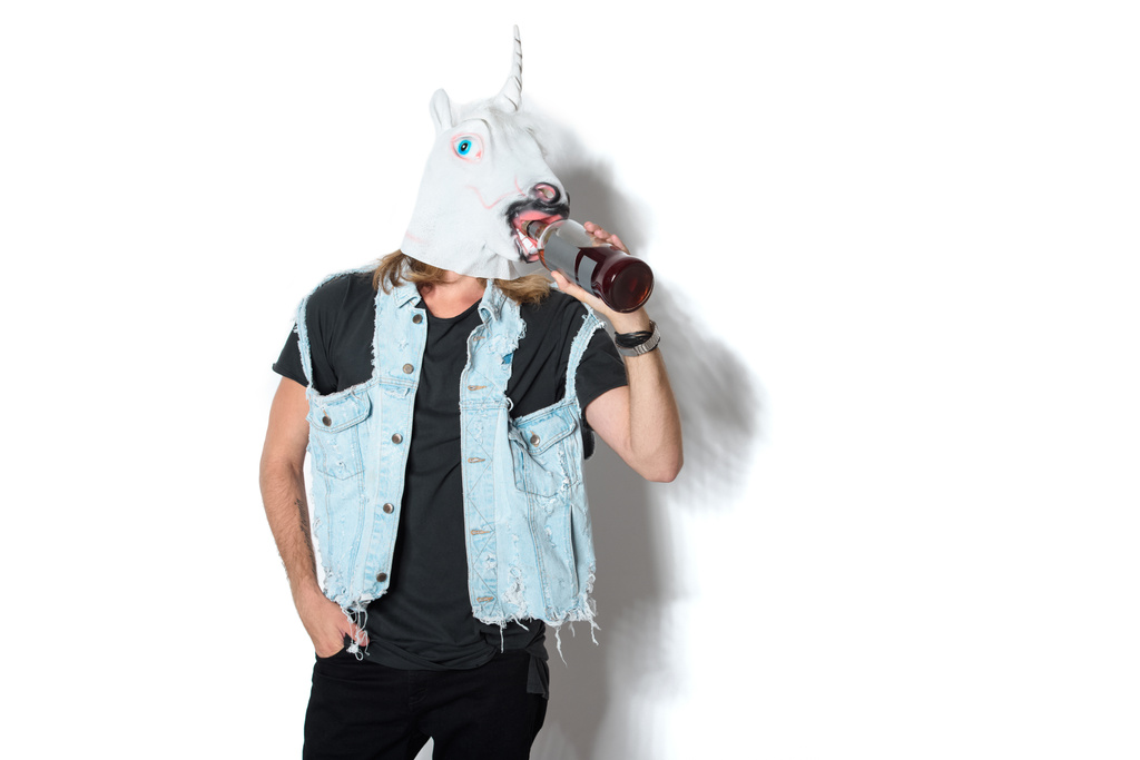man in unicorn mask and denim vest drinking rum through mask on white - Photo, Image