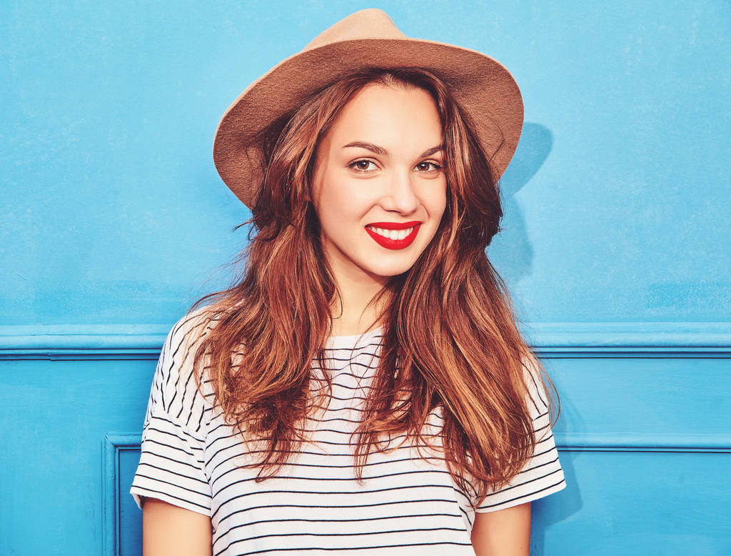 Νεαρό όμορφο κορίτσι μοντέλο σε καφετί καπέλο με κόκκινα χείλη, θέτοντας κοντά μπλε τοίχο και casual καλοκαίρι ρούχα - Φωτογραφία, εικόνα