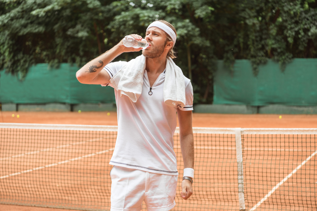 Retro gestylte müde Tennisspielerin mit Handtuch trinkt Wasser auf Tennisplatz - Foto, Bild