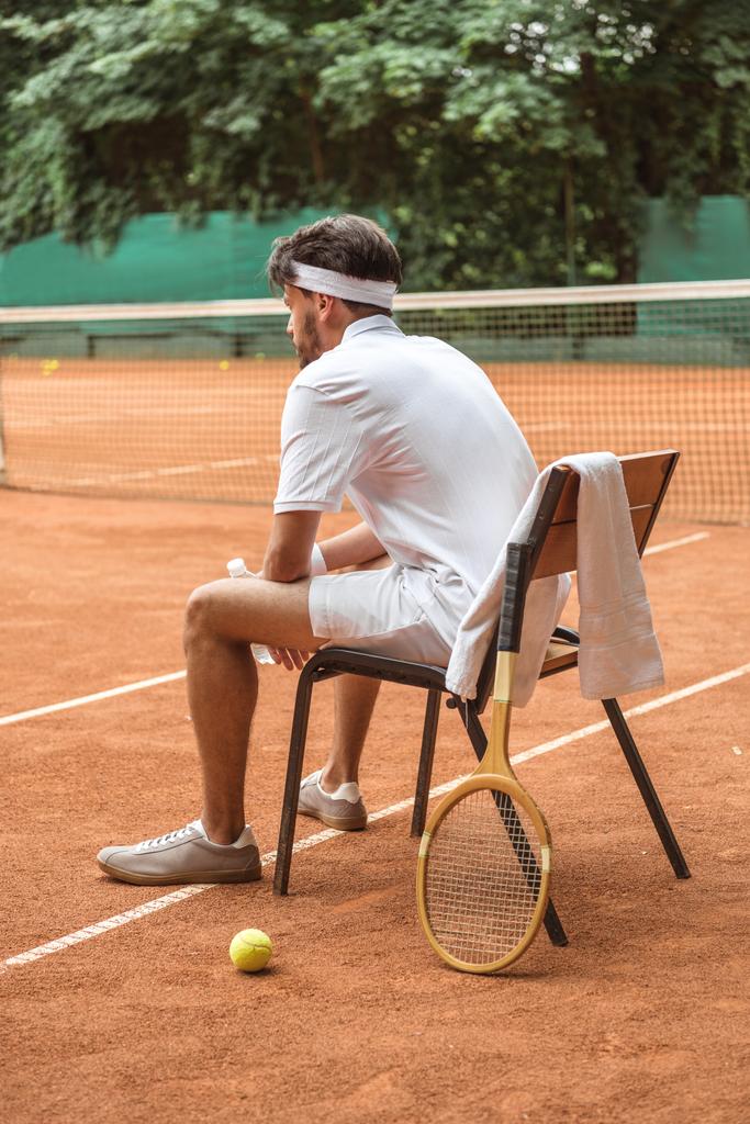 παίκτης του τένις στο λευκό sportswear ακουμπά στην καρέκλα με μπάλα του τένις, ρετρό ξύλινη ρακέτα και πετσέτα σε δικαστήριο - Φωτογραφία, εικόνα