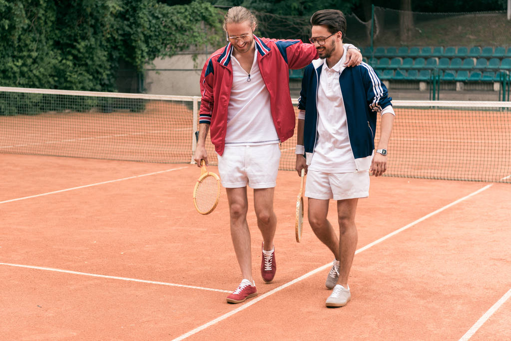 fröhliche sportliche Freunde mit Holzschlägern umarmen und auf dem Tennisplatz gehen - Foto, Bild