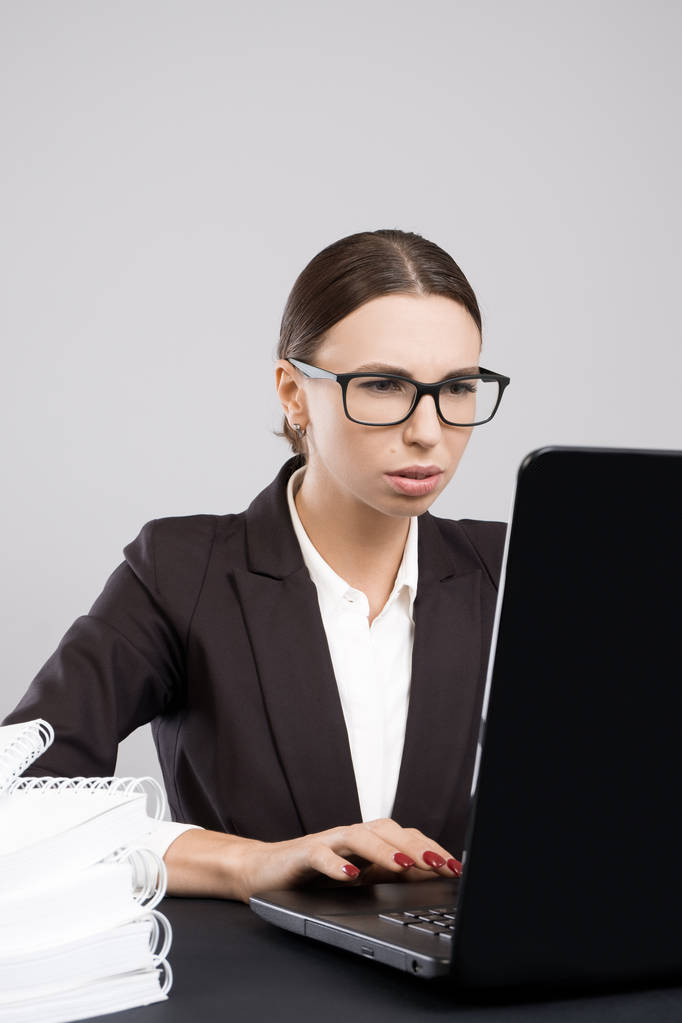 junge aufmerksame Geschäftsfrau sitzt mit Papieren und Büchern am Schreibtisch und benutzt Laptop - Foto, Bild