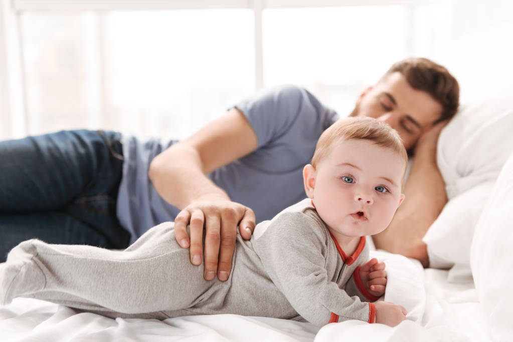 Image de beau jeune homme père papa dormant au lit tandis que son petit enfant fils regardant caméra à l'intérieur à la maison
. - Photo, image