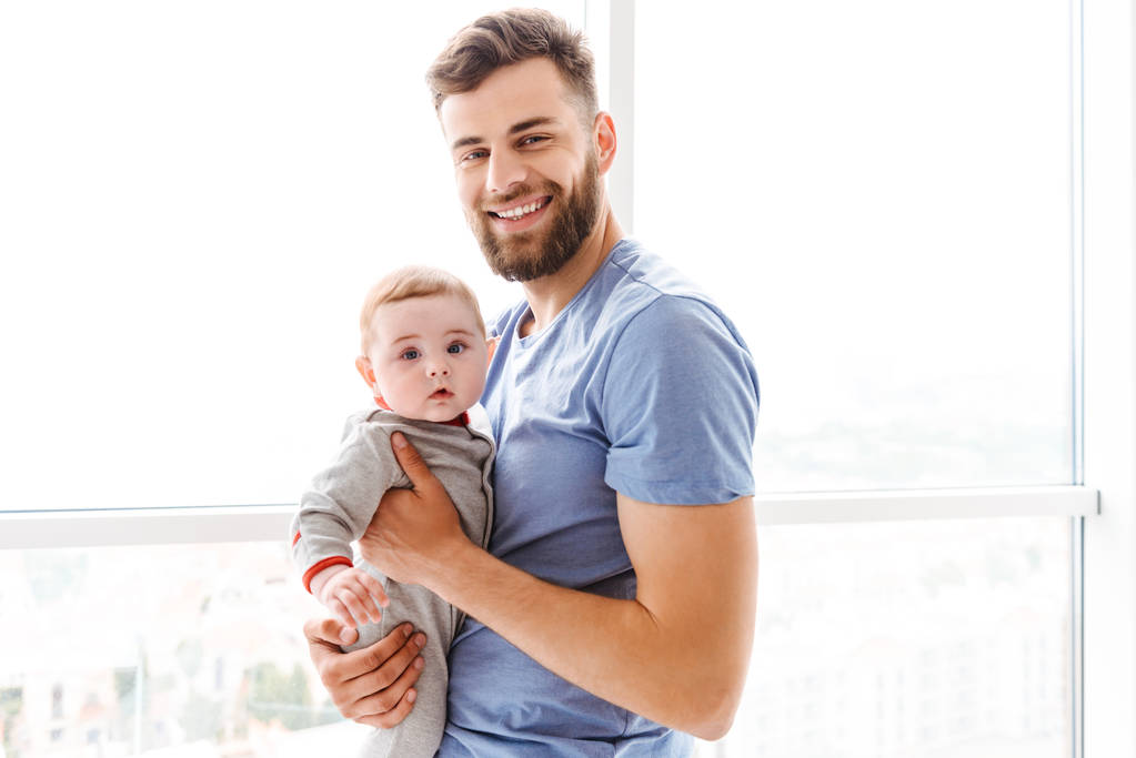 Obrázek pohledný mladý vousatý muž drží jeho malý syn dítě v rukou pohledu kamery. - Fotografie, Obrázek