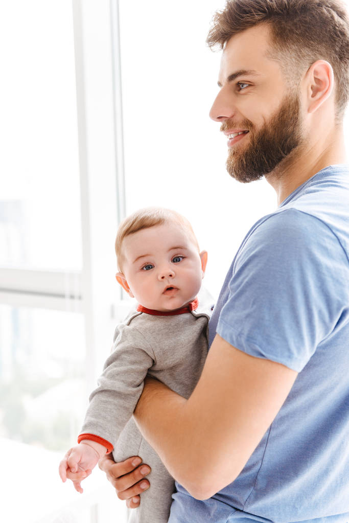 Image of handsome young cheerful bearded man holding his little son child in hands looking aside. - Photo, Image