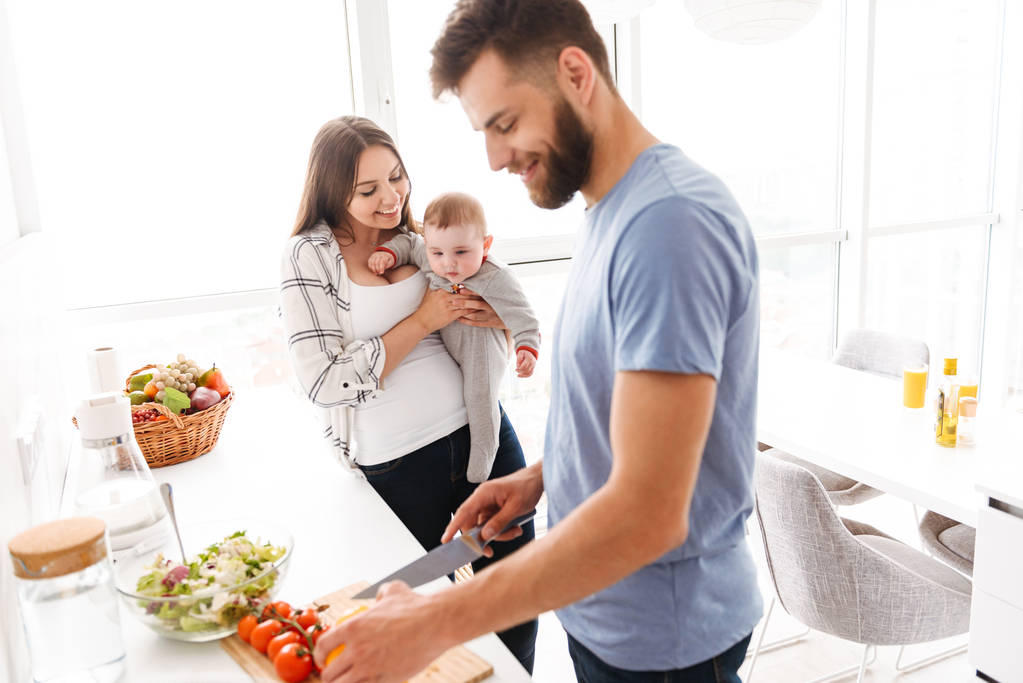 キッチンで料理をして小さな子供赤ちゃん息子と一緒にいくつかの両親を愛する幸せな若者のイメージ. - 写真・画像