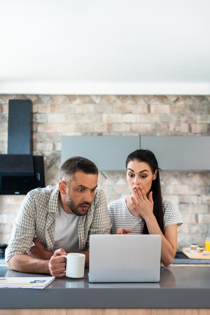 портрет шокированной супружеской пары, смотрящей вместе на экран ноутбука на кухне
 - Фото, изображение