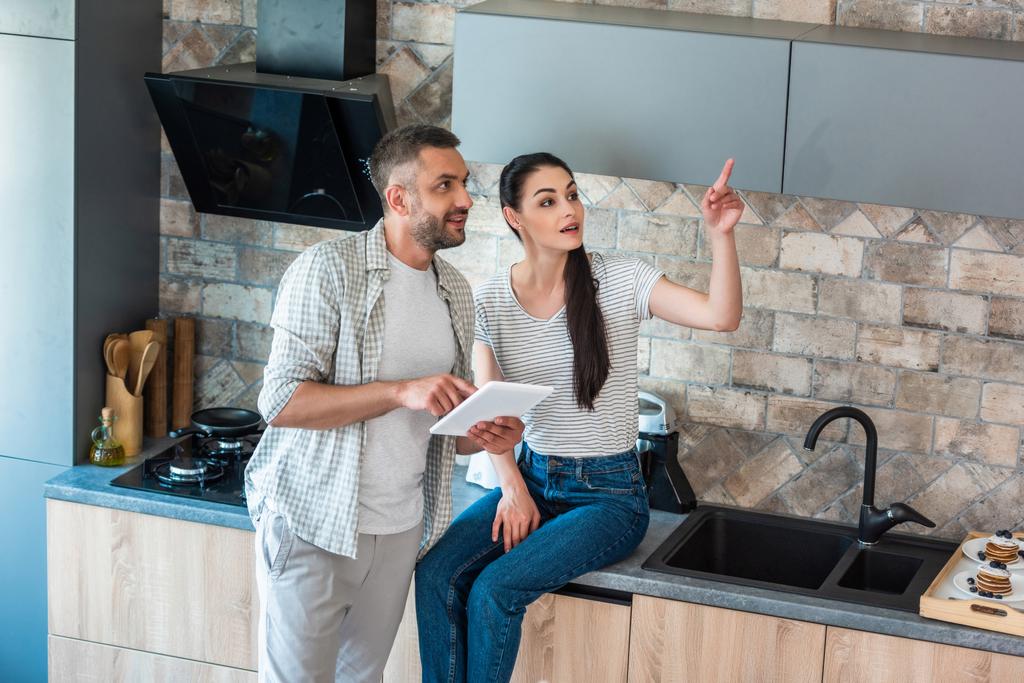 married couple with digital tablet looking away in kitchen, smart home concept - Photo, Image